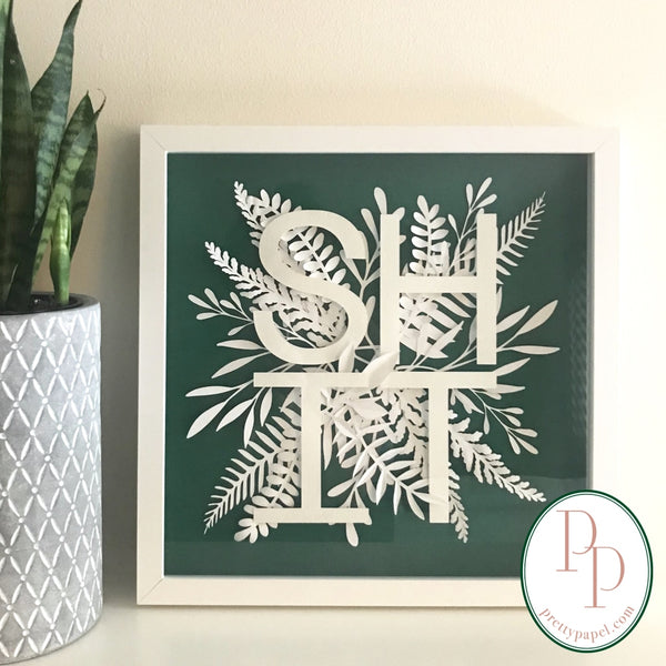 Botanical paper cut collage with woodland foliage and clean, sans serif letters spelling SHIT on top of a bright green background. In white square shadowbox frame.