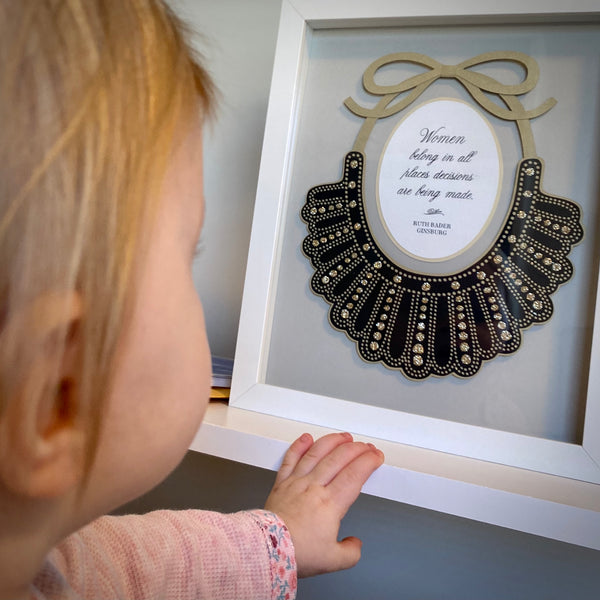 A little girl reaches out to framed, papercut artwork of Ruth Bader Ginsburg's iconic Dissent Collar. Quote inside reads, "Women belong in all places decisions are being made."
