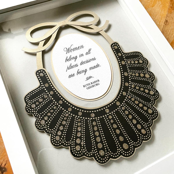 Papercut artwork of Ruth Bader Ginsburg's iconic Dissent Collar, framed in a white shadowbox. Quote inside reads, "Women belong in all places decisions are being made."